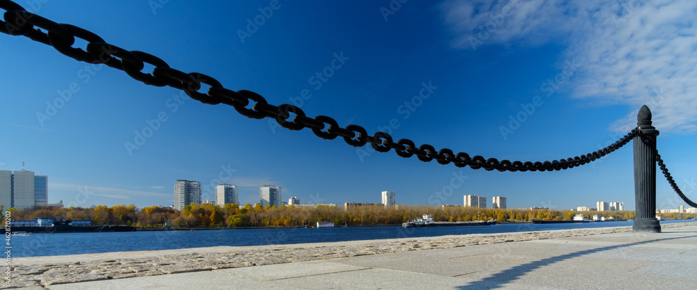 Fototapeta premium A fragment of a cast-iron chain fence on the river embankment. In the background, water, blue skies and high-rise buildings on the opposite bank of the river. Sunny day. Panorama.