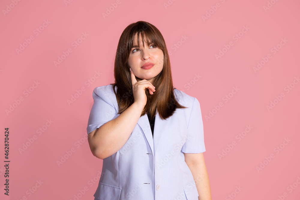 Young woman wearing classic business clothes on pink background put her hand to face, looks up, thoughtful portrait in studio on pink background. Young woman wearing classic business clothes on pink background put her hand to face, looks up, thoughtful portrait in studio on pink background.