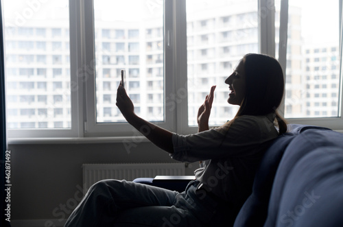 Silhouette of smiling woman sitting down on the comfortable couch and talking on the smartphone via video connect, cheerful girl waving, speaking with friends, using mobile app for virtual meeting