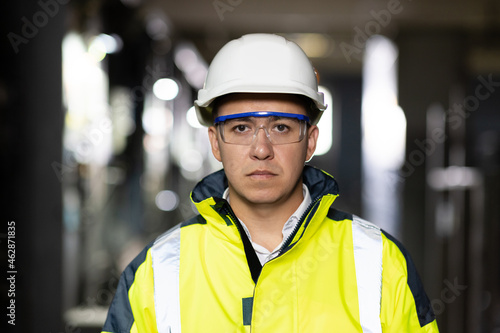 Portrait of employee serious asian man engineer worker wearing safety uniform, goggles and hardhat looking at camera on site factory warehouse background