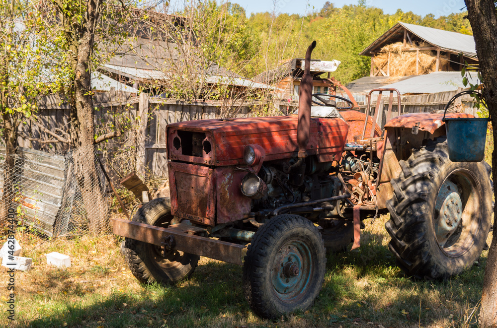Old red tractor parked on the green grass in the backyard of the country house. Tractor in the farm yard, autumn landscape background Old red tractor parked on the green grass in the backyard of the country house. Tractor in the farm yard, autumn landscape background