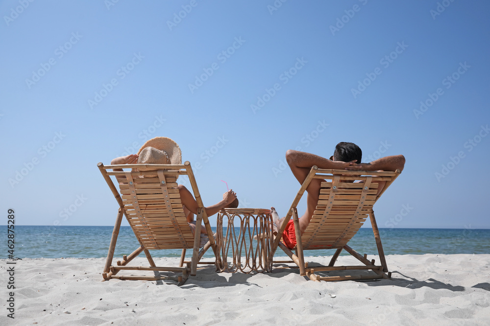 Couple resting in wooden sunbeds on tropical beach - obrazy, fototapety, plakaty 