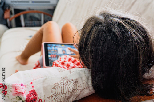 Child lying on sofa and using tablet computer, concept of using tablet in distance education. Cute little girl playing game, studying or watching cartoon on digital device.	 photo