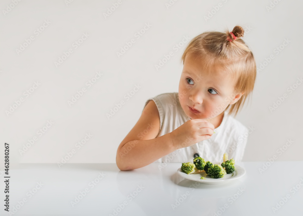 Cute funny little toddler girl eating broccoli at the table, white walls. Childhood, healthy food and children's emotions Cute funny little toddler girl eating broccoli at the table, white walls. Childhood, healthy food and children's emotions