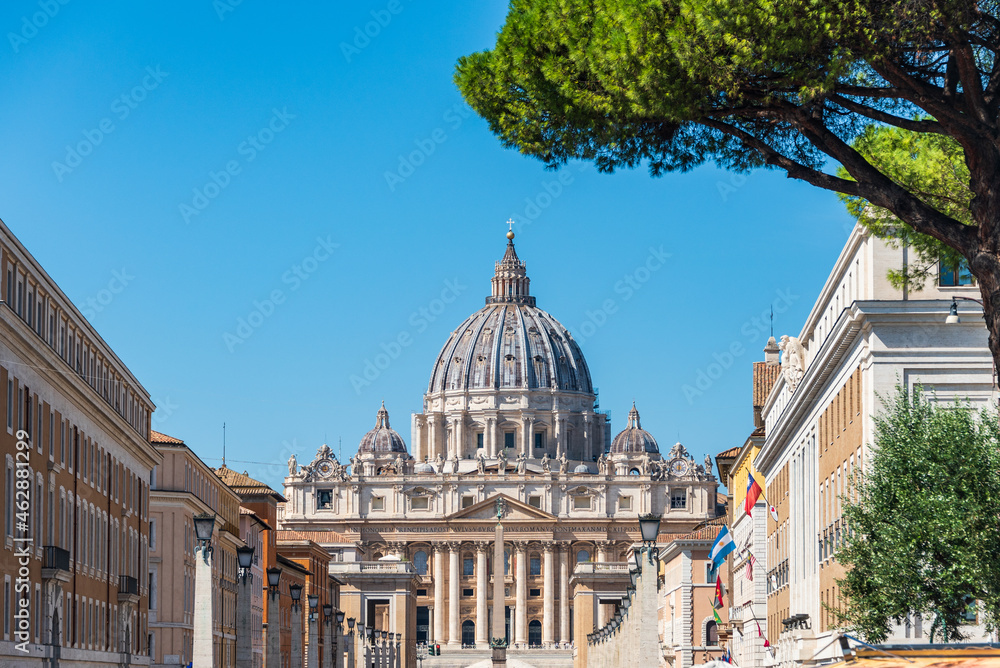 The dome of Saint Peter's Basilica and its statues of saints at St. Peter's Square, Vatican City view from Via della Conciliazione in Rome, Italy. Pine tree at the foreground.