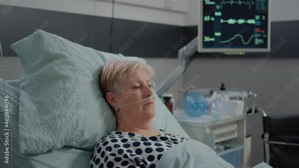 Portrait of elder patient with oxygen tube laying in hospital ward bed. Woman with illness looking at camera while waiting for healthcare assistance in emergency room for intensive care Portrait of elder patient with oxygen tube laying in hospital ward bed. Woman with illness looking at camera while waiting for healthcare assistance in emergency room for intensive care
