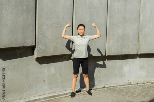 Happy brunette Asian sportswoman in light t-shirt shows arm muscles standing by wall with concrete plates on street