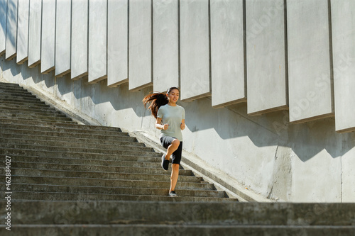 Asian woman in tracksuit runs down underground crossing large stone stairs past wall with panels training on city street in summer morning