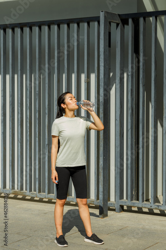 Fototapeta Naklejka Na Ścianę i Meble -  Athletic Asian lady with ponytail drinks water from plastic bottle resting after training on sunny city street