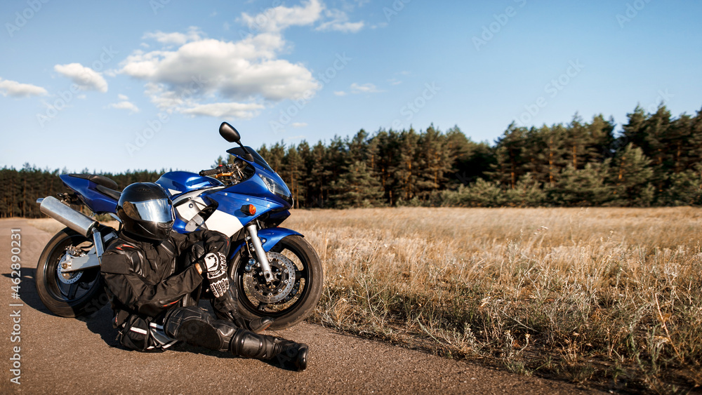 Motorcycle driver in helmet and leather jacket sits on the asphalt road next to a sports motorcycle Motorcycle driver in helmet and leather jacket sits on the asphalt road next to a sports motorcycle