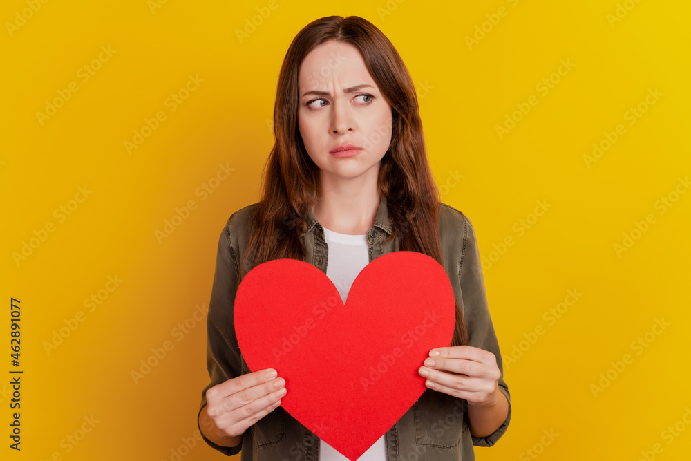 Portrait of upset unsure girl hold red paper heart shape symbol look empty space on yellow background Portrait of upset unsure girl hold red paper heart shape symbol look empty space on yellow background
