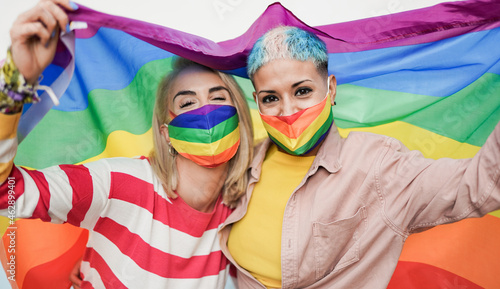 Happy lesbian women with lgbt rainbow flag at gay pride parade wearing colorful safety face mask for coronavirus outbreak