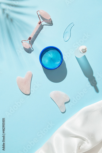 Cosmetic facial kit for home skin care and spa. Collagen eye patches, face roller and guasha massagers, bottle of essential oils or serum on blue pastel background. Top view. Flat lay