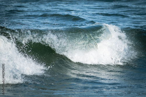 An angry turquoise green color massive rip curl of a wave as it rolls along a beach. The white mist and froth from the wave are foamy and fluffy. The Atlantic Ocean in the background is deep blue. 