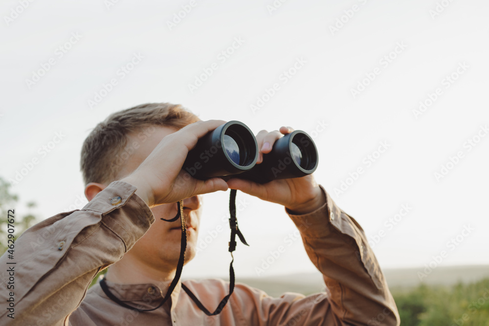 A young man looks through binoculars at a beautiful hilly landscape.The concept of hunting, travel and outdoor recreation. Banner with copy space.A traveler or hunter is observing through binoculars. A young man looks through binoculars at a beautiful hilly landscape.The concept of hunting, travel and outdoor recreation. Banner with copy space.A traveler or hunter is observing through binoculars.