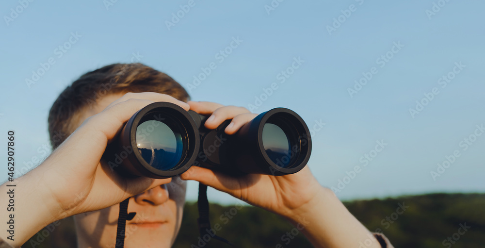 A man with modern binoculars against the sky and green forest.The concept of hunting, travel and outdoor recreation. Banner with copy space.A traveler or hunter is observing through binoculars. A man with modern binoculars against the sky and green forest.The concept of hunting, travel and outdoor recreation. Banner with copy space.A traveler or hunter is observing through binoculars.