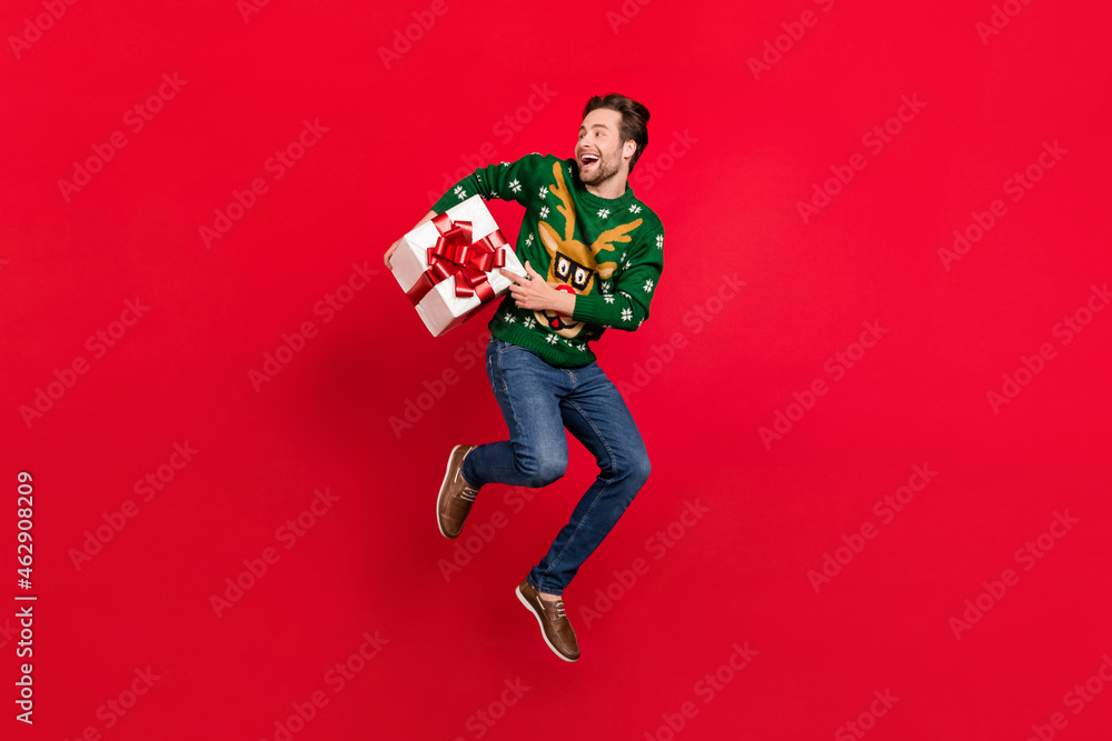 Full size photo of happy smiling good mood guy in ugly sweater jump hold present box isolated on red color background Full size photo of happy smiling good mood guy in ugly sweater jump hold present box isolated on red color background