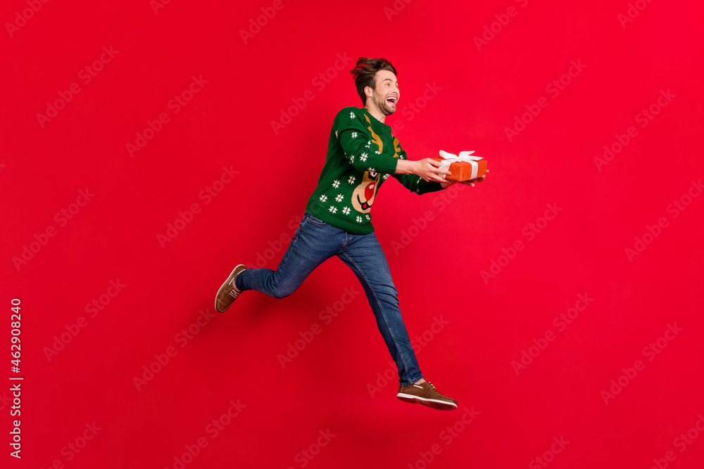 Full size profile side photo of smiling crazy running man give christmas present celebration isolated on red color background Full size profile side photo of smiling crazy running man give christmas present celebration isolated on red color background
