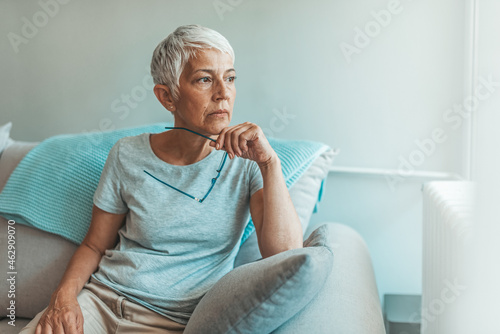 Upset depressed senior gray-haired lady touching temple, sitting on sofa, looking in the window, feeling worried and stressed about the future and thinking about own problems photo