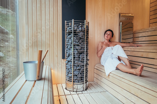 Foto Relaxing Woman wrapped white towel lying on the wooden bench in Hot Finnish sauna with a wide window near hot stones and enjoying pleasant body care temperature treatment
