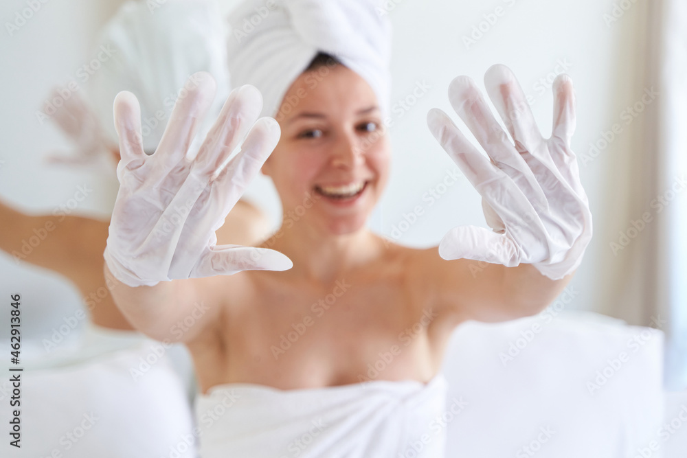 After a shower, a girl wrapped in a towel uses cosmetic gloves to moisturize the skin of her hands. Cosmetic trends for body care at home