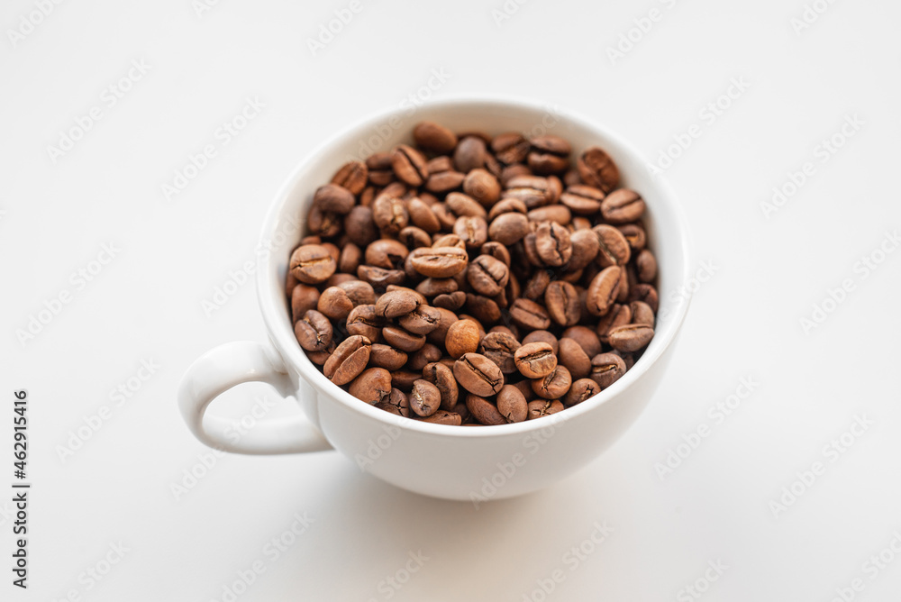 A white cup filled with brown coffee beans on white background