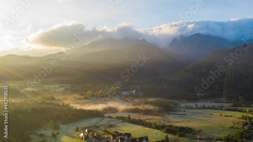 Aerial time lapse hyperlapse foggy morning in alpine village lit with warm sunlight. Drone shot sun rises over epic mountain peaks covered with rolling clouds. Incredible view valley in sunrise light