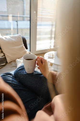A cup of tea or coffee in the hands of a girl. In the morning, the girl drinks hot tea. Enjoy the comforts of home. White cup close-up in the hands of a woman in the rays of the sun from the window