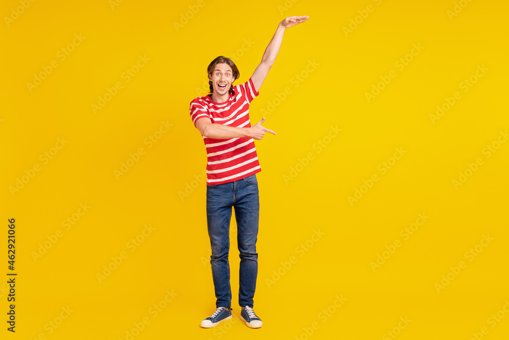 Photo of sweet impressed young man wear striped t-shirt measuring size pointing finger empty space isolated yellow color background Photo of sweet impressed young man wear striped t-shirt measuring size pointing finger empty space isolated yellow color background