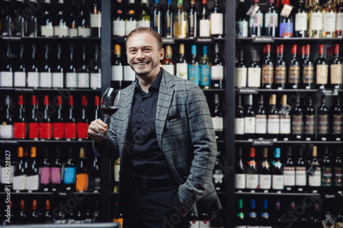 Portrait of happy man with glass of red wine on background of showcase of alcohol store