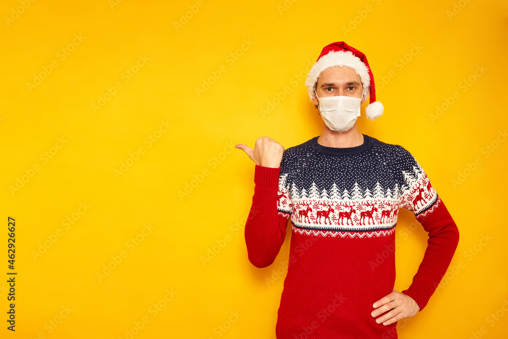 man in Christmas sweater, medical mask, red Santa Claus hat points with his index finger at an empty area for New Year advertising. isolated yellow background with copy space concept - people, holiday