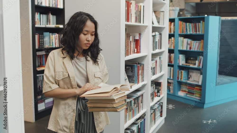 Medium slowmo shot of female Asian university student standing among bookshelves at contemporary library holding small pile of books reading one Medium slowmo shot of female Asian university student standing among bookshelves at contemporary library holding small pile of books reading one