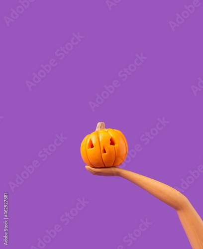 One pumpkin in a plastic doll's hand on a pastel purple background Minimalistic Halloween concept. Creative idea.