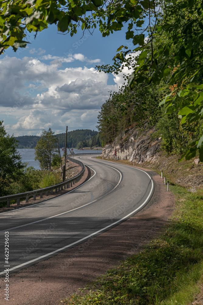 Curve of an empty asphalt countryside highway. The road has 2 lanes, fenced and with white markings. Large stones, rocks and trees is on the side of the road, on the other side there is a Ladoga lake.