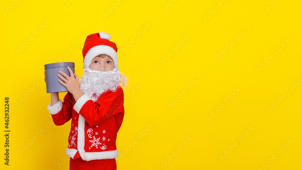 Little boy in Santa Claus clothes and with an artificial beard holding a round gift box in his hands, his gaze is directed to the side, yellow, copy space. Little boy in Santa Claus clothes and with an artificial beard holding a round gift box in his hands, his gaze is directed to the side, yellow, copy space.