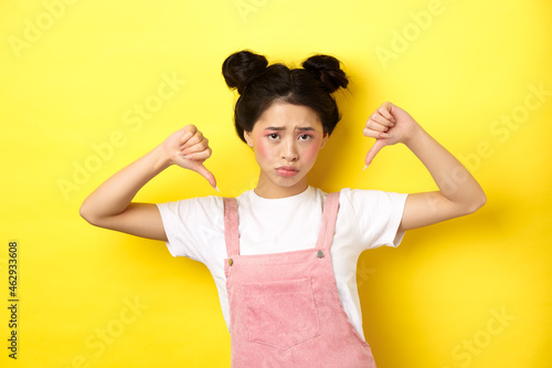 Summer lifestyle concept. Sad and gloomy asian woman with makeup, pointing fingers down and frowning upset, disappointed with unfair situation, yellow background photo