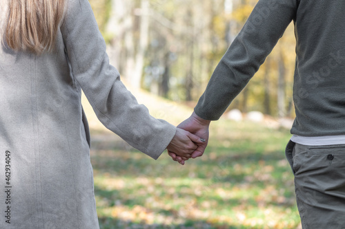 Love, relationship, nature concept. Close-up shot from back of man and woman holds each other hands in blurred nature background during cold and sunny day