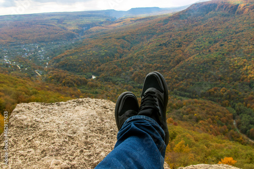 Legs of a hiker in sneakers sitting on the edge of a cliff and looking at the forest and mountains