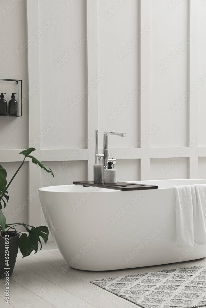 Fototapeta premium Modern ceramic bathtub and green plant near white wall indoors