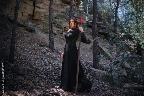 Evil witch with a staff in the forest. Slavic witch creates red magic. A young woman in a black dress and a crown embroidered with beads.