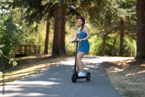 An environmentally friendly form of transport - an electric scooter. A young girl in a protective helmet and denim dress rides in the park. Courier, outdoor activities, sports.