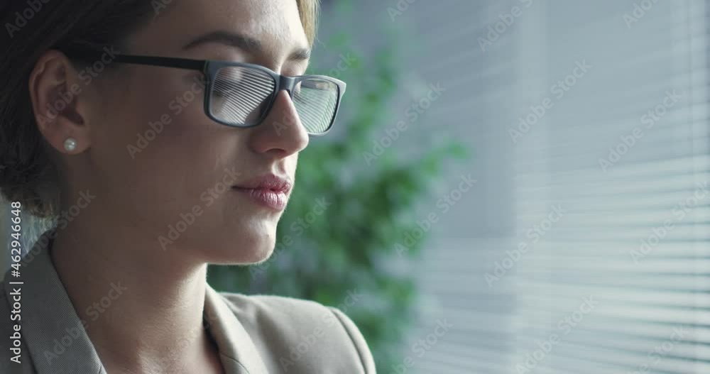 Close up of beautiful caucasian woman with glasses sitting in office and looking thoughtfully out the window thinking about work task.