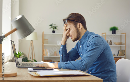 Tired worker having problem. Man makes dumb mistake or has difficulty with digital file. Side view of upset stressed young guy facepalming sitting at working desk with laptop computer in home office photo