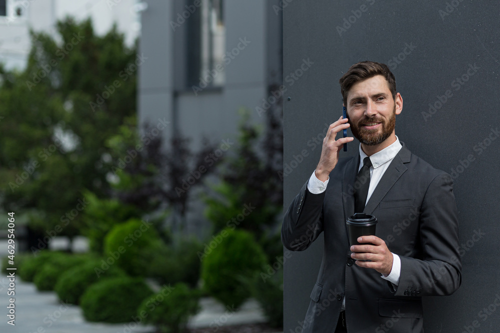 Successful businessman broker in a business suit talking on the phone near the office outside, Banker holding a cup of coffee during lunch break smiling and looking at camera Successful businessman broker in a business suit talking on the phone near the office outside, Banker holding a cup of coffee during lunch break smiling and looking at camera