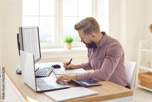 Serious young businessman or financial accountant sitting in office workplace at desk with desktop and laptop computers, writing something, doing multiple tasks, working with data files, taking notes