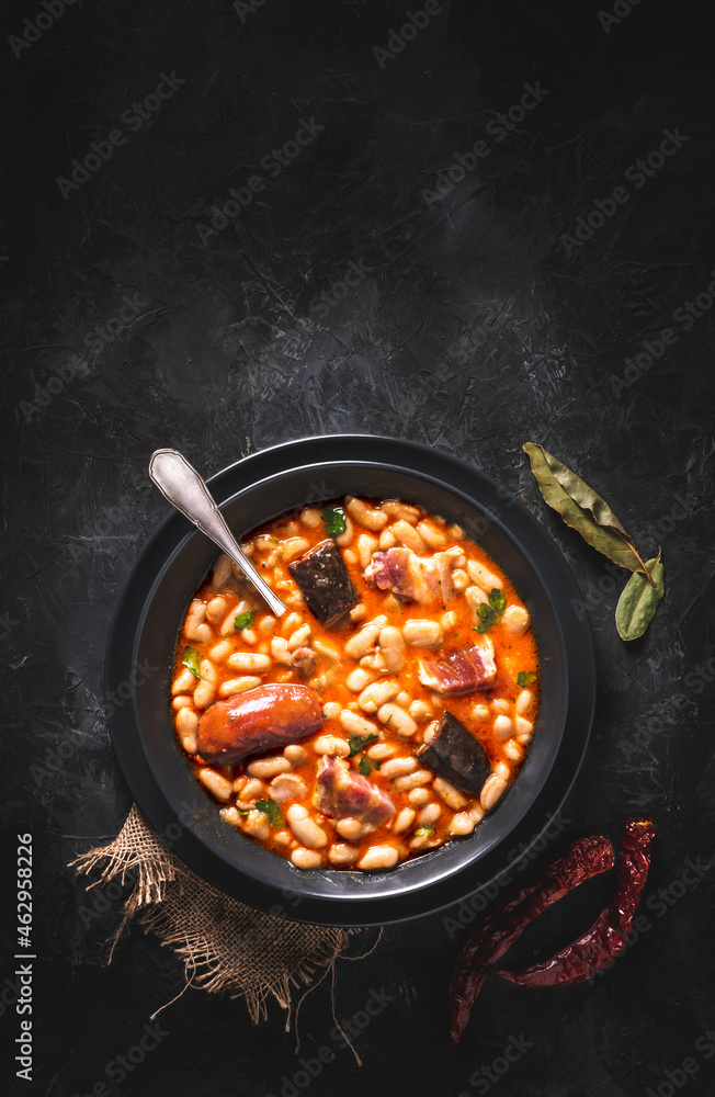 Traditional recipe of beans with chorizo and blood sausage called “Fabada Asturiana” on a dark table with some ingredients around. Typical Spanish food with “compango”. 