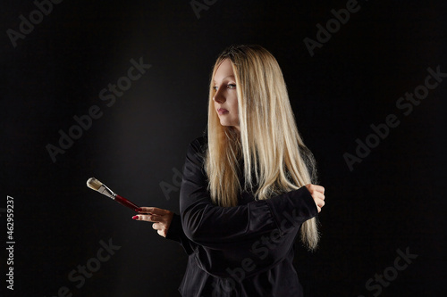 young blonde artist poses with a brush in her hand. photo shoot on a black background in the studio