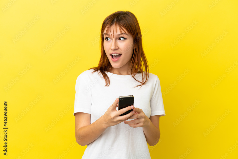 Redhead girl isolated on yellow background using mobile phone and looking up Redhead girl isolated on yellow background using mobile phone and looking up
