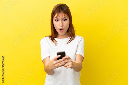 Redhead girl isolated on yellow background looking at the camera while using the mobile with surprised expression