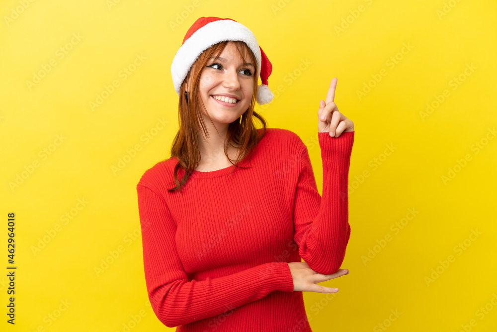 Redhead girl with christmas hat isolated on yellow background pointing up a great idea Redhead girl with christmas hat isolated on yellow background pointing up a great idea